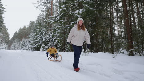 happy-family-in-snowy-forest-in-weekend-mother-is-pulling-sledge-with-little-child-woman-and-child